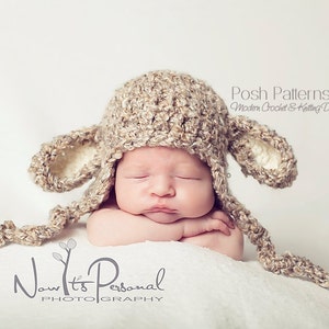 May include: A newborn baby wearing a crocheted brown and white lamb hat with long ear flaps. The hat has a long tail that is tied under the chin. The baby is sleeping peacefully with their arms crossed in front of them. The background is a white surface.