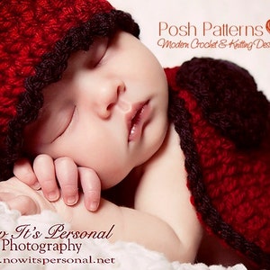 May include: A red crocheted baby hat and matching cape with black trim. The hat has a scalloped edge. The baby is sleeping peacefully.
