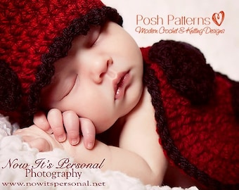 May include: A red crocheted baby hat and matching cape with black trim. The hat has a scalloped edge. The baby is sleeping peacefully.