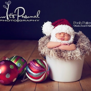 Puede incluir: Un beb&eacute; reci&eacute;n nacido lleva un gorro de Pap&aacute; Noel de crochet blanco y rojo con un pomp&oacute;n blanco y duerme en un cubo blanco forrado de piel sint&eacute;tica marr&oacute;n. Tres adornos navide&ntilde;os en rojo, verde y blanco est&aacute;n en primer plano.
