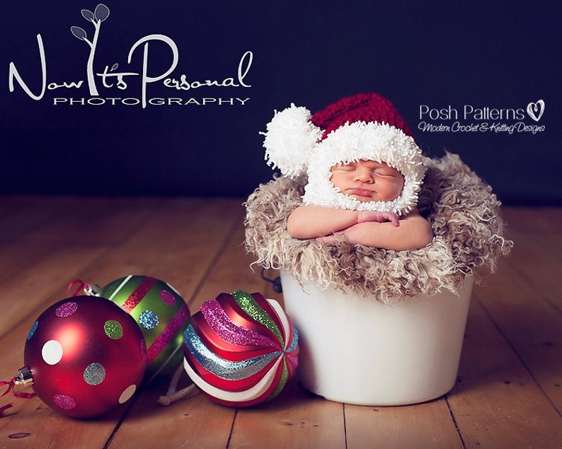 Puede incluir: Un beb&eacute; reci&eacute;n nacido lleva un gorro de Pap&aacute; Noel de crochet blanco y rojo con un pomp&oacute;n blanco y duerme en un cubo blanco forrado de piel sint&eacute;tica marr&oacute;n. Tres adornos navide&ntilde;os en rojo, verde y blanco est&aacute;n en primer plano.