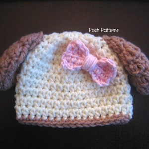 May include: A crocheted baby hat with brown ear flaps and a pink bow. The hat is white with a brown trim. The text "Posh Patterns" is visible on the hat.
