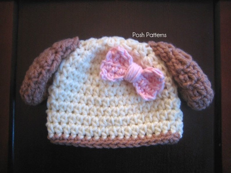 May include: A crocheted baby hat with brown ear flaps and a pink bow. The hat is white with a brown trim. The text "Posh Patterns" is visible on the hat.