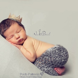May include: A newborn baby wearing a gold lace crown and a gray crocheted diaper cover, sleeping on a white pillow.