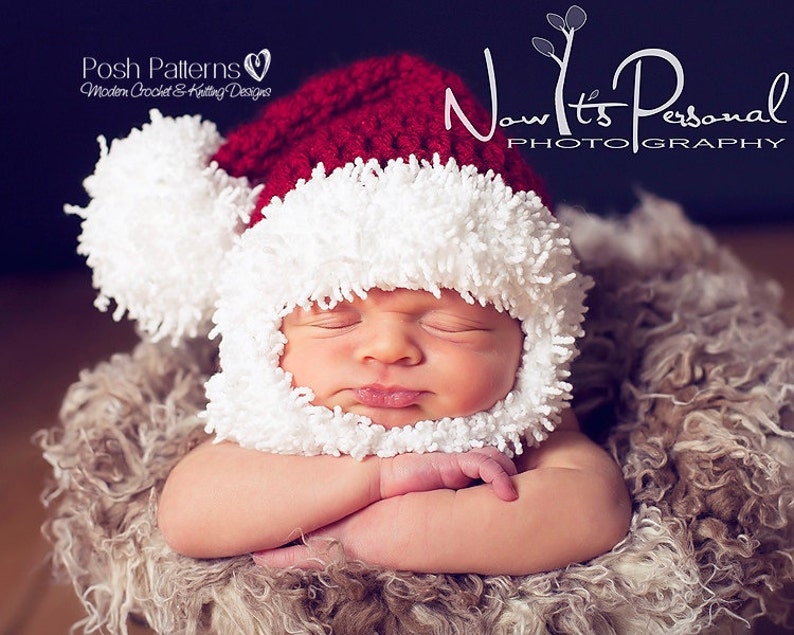 Puede incluir: Un beb&eacute; reci&eacute;n nacido con un gorro de Pap&aacute; Noel de crochet blanco y rojo con un pomp&oacute;n esponjoso. El beb&eacute; est&aacute; durmiendo con los brazos cruzados y est&aacute; acostado sobre una superficie esponjosa marr&oacute;n.