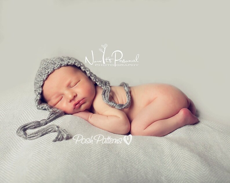 May include: A newborn baby wearing a gray knitted hat with a long tassel sleeps on a white blanket. The baby is curled up with their arms and legs tucked in. The text "Now It's Personal Photography" and "Posh Patterns" is visible in the image.
