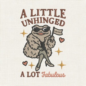 Op de afbeelding: Geborduurd ontwerp met een kikker met zonnebril en een bontjas, met een vlag met de tekst "Thriving". De tekst hierboven luidt "A LITTLE UNHINGED" en hieronder "A LOT Fabulous". Het ontwerp bevat harten en sterren.