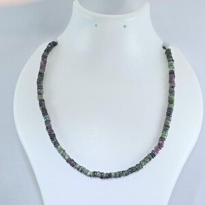 May include: A necklace composed of small, square-cut gemstones in shades of green, purple, and black. The necklace is displayed on a white mannequin bust, showcasing the jewelry's design and color variations. The gemstones are evenly spaced.
