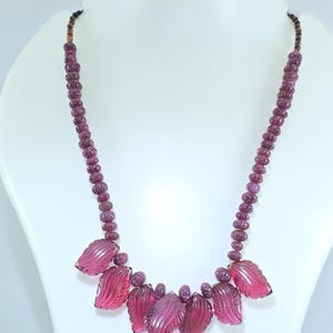 May include: A necklace featuring a series of small, round, dark pink beads. The necklace has a series of carved leaf-shaped pendants in a lighter shade of pink. The necklace is displayed on a white mannequin.