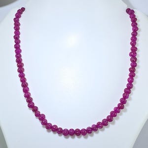 Può includere: Una collana con piccole perline rotonde di colore magenta. Le perline hanno una texture a coste e sono infilate insieme per formare una collana completa. La collana è esposta su un espositore di gioielli bianco.