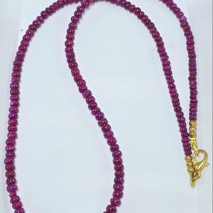 May include: A necklace composed of small, round, deep pink gemstones. The beads are strung together, forming a continuous loop. The clasp and connecting elements are gold-colored. The necklace is displayed against a white background.