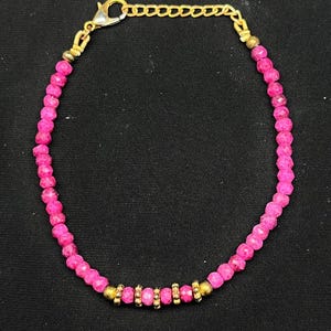 May include: A pink beaded bracelet with gold-colored accents and a clasp. The bracelet features small, faceted pink beads strung together with gold-colored spacers and a gold-colored chain for adjusting the size. The clasp is also gold-colored.