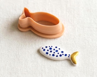 May include: A peach-colored plastic fish-shaped cookie cutter positioned above a small white ceramic fish figurine featuring blue polka dots and a gold-painted tail fin, both displayed on a textured off-white fabric surface.