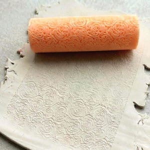 Könnte beinhalten: Eine orangefarbene Walze mit Rosenmuster liegt auf einem Tonstück, das ebenfalls mit einem Rosenmuster bedruckt ist. Der Ton ist cremefarben und hat einen strukturierten, unebenen Rand. Die Walze ist zylindrisch.