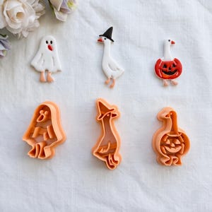Peut inclure: Trois figurines en argile blanche représentant un fantôme, une oie avec un chapeau noir et une oie en costume de citrouille, accompagnées de trois emporte-pièces en plastique orange assortis avec des détails noirs, rouges et orange.