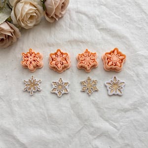May include: Four peach-coloured plastic snowflake cookie cutters arranged in a row above four corresponding white and gold snowflake-shaped fondant or cookie cutouts, each featuring intricate geometric patterns and detailed embossed designs.