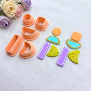 May include: A set of peach-colored plastic clay cutters in various geometric shapes including rectangles, squares, and semi-circles, alongside finished clay pieces in orange, blue, purple, and lime green with painted details.