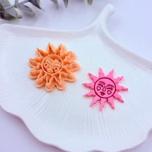 Peut inclure: Deux emporte-pièces en forme de soleil. L'un est orange, l'autre rose. Les deux ont un visage au centre et des bords en forme de rayons. Les emporte-pièces sont présentés sur une assiette blanche en forme de feuille.