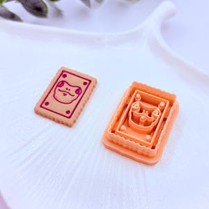 Könnte beinhalten: Ein tan-farbener Keks mit einem Froschgesicht-Design und einem passenden Ausstecher. Der Keks hat eine rechteckige Form mit einem gewellten Rand und einem rosa Froschgesicht-Design. Der Ausstecher ist orange.