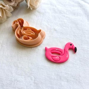 May include: A pink flamingo-shaped cookie cutter and a pink flamingo-shaped decoration are displayed on a white surface. The cookie cutter is a light peach color. The flamingo decoration has a black beak.