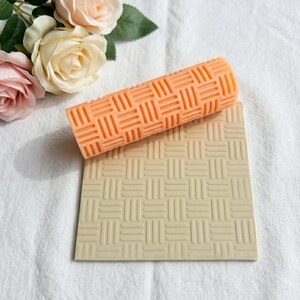 May include: A cylindrical orange rolling pin with a raised basketweave pattern and a square beige mat featuring the same embossed basketweave texture, accompanied by three pale pink and cream-coloured artificial roses with green leaves.