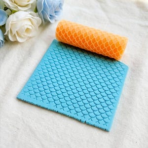 May include: A blue silicone mat and orange rolling pin, both with a mermaid scale design. The rectangular mat and cylindrical rolling pin are for baking. The items are designed for decorating cakes and cookies.