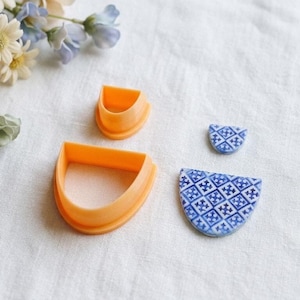 Könnte beinhalten: Draufsicht auf zwei orangefarbene Polymer Clay Cutter in Halbkreisform, neben zwei blau-weiß gemusterten Keramikteilen. Die Keramikteile haben ein geometrisches Design. Der Hintergrund ist eine weiße Oberfläche.