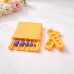 May include: An orange bead reamer set with a sliding lid, containing five purple beads. Two additional orange bead measuring tools are also present. The tools are designed for jewelry making and beadwork.