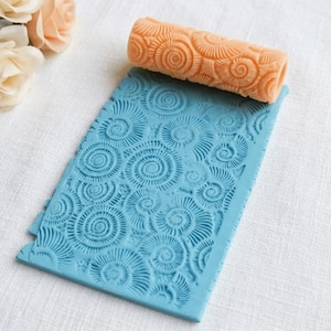 May include: A light orange textured rolling pin sits on a blue rectangular mat, both with a spiral design. The rolling pin is cylindrical, and the mat has a repeating spiral pattern. A bouquet of peach-coloured roses is in the background.