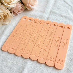 May include: A collection of peach-colored, rectangular tools with inspirational phrases like "Be yourself" and "Trust your art." Each tool has a different measurement in millimeters and a decorative top, such as a star or a crown.