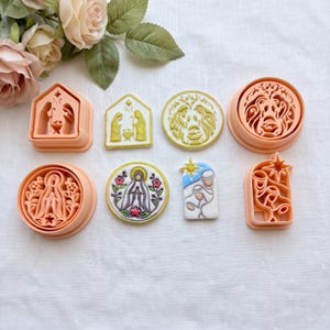 May include: A collection of peach-colored plastic cookie cutters and matching decorated cookies featuring religious nativity and floral motifs. The set includes house-shaped, circular, and arched designs with intricate embossed details of figures and stars.
