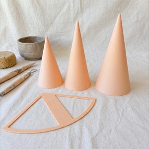 May include: Three smooth, light peach-colored cones of varying heights, a flat peach-colored cone-shaped template, two wooden sculpting tools, a small gray ceramic bowl, and a round, textured tan sponge are arranged together.