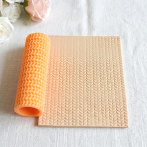 Könnte beinhalten: Eine orangefarbene Silikon-Teigrolle und eine beige strukturierte Matte mit einem Blättermuster. Die Teigrolle ist zylindrisch mit einer strukturierten Oberfläche. Die Matte ist rechteckig und hat ein sich wiederholendes Blattdesign, geeignet zum Backen und Basteln.
