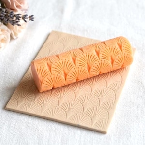 May include: An orange patterned roller rests on a beige, rectangular, patterned surface. The roller and surface feature a repeating fan-like design. The items are on a white surface, with flowers in the background.