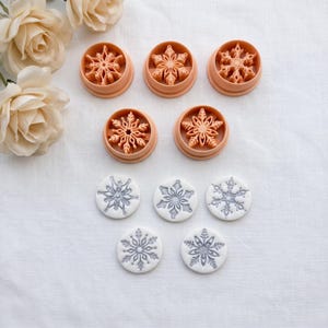 May include: Five peach-colored circular cookie cutters featuring intricate snowflake designs, accompanied by five corresponding white circular clay or dough impressions that display the detailed snowflake patterns in a light gray color.