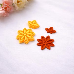 Könnte beinhalten: Vier blütenförmige Ausstecher in Gelb- und Orangetönen sind auf einer weißen Oberfläche angeordnet. Die Ausstecher variieren in Größe und Design, mit einer großen gelben Blume und einer kleineren orangefarbenen Blume. Backwerkzeuge mit floralem Thema.