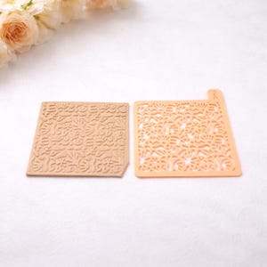 May include: Two craft tools: a tan embossed square and a peach-coloured stencil. The embossed square has a repeating floral pattern. The stencil has a similar floral design, with the pattern cut out. Both are on a white surface.