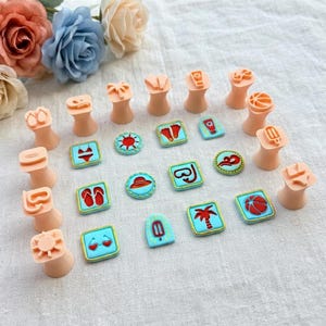 May include: A collection of beach-themed cookie stamps and matching cookies. The stamps are peach-colored and feature designs like a sun, flip-flops, and a bikini. The cookies are square or round with blue frosting and red and yellow details.