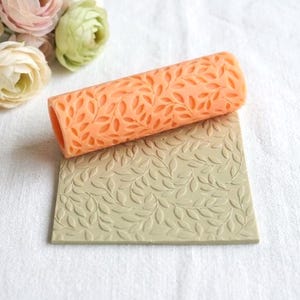 May include: An orange leaf-patterned rolling pin rests on a rectangular, leaf-patterned, light green surface. The rolling pin is cylindrical and has a hollow center. The background includes soft, light-colored flowers.