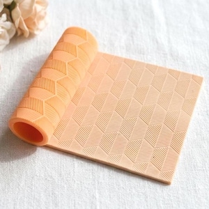 May include: A rolled-up, peach-colored silicone mat with a geometric pattern. The mat is partially unrolled, revealing a textured surface with a repeating design of angled lines and shapes. The mat is designed for kitchen use.