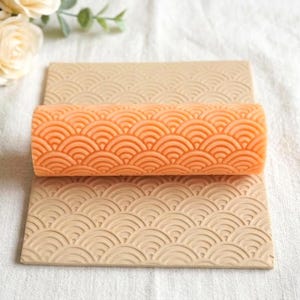 Könnte beinhalten: Eine orangefarbene, gemusterte Teigrolle liegt auf zwei beigefarbenen, strukturierten rechteckigen Brettern. Die Teigrolle und die Bretter weisen ein sich wiederholendes wellenartiges Design auf. Die Artikel werden wahrscheinlich zum Backen oder Basteln verwendet.