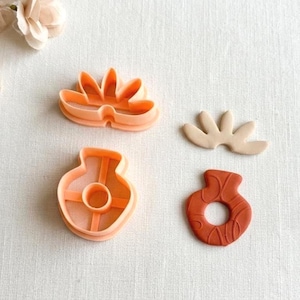 Puede incluir: Cuatro elementos son visibles: dos cortadores de galletas de plástico naranja, uno con forma de flor y otro con forma de jarrón. También hay dos galletas, una con forma de flor y otra con forma de jarrón, en colores beige y marrón.