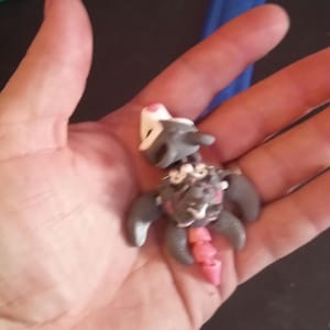 May include: A handmade clay figurine of a creature with gray, white, and pink details. The figurine has a small head, large wings, and a long, segmented tail. The figure is held in a hand.
