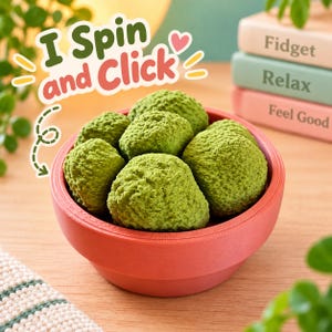May include: A round, coral-colored bowl filled with several textured, bright green, spherical fidget objects. The text "I Spin and Click" is printed in green and red above the bowl, accompanied by a small pink heart and yellow dashes.
