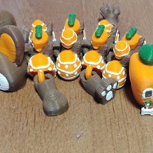 May include: A collection of 3D-printed miniature figurines, including a brown deer with orange ears, several orange mushrooms with white spots, and a small orange house with a green top and windows. The figurines are on a wooden surface.