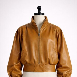 Peut inclure: Veste de bombardier en cuir marron clair avec fermeture éclair, col montant et poignets et taille élastiqués. La veste a deux poches avant et est présentée sur un mannequin.