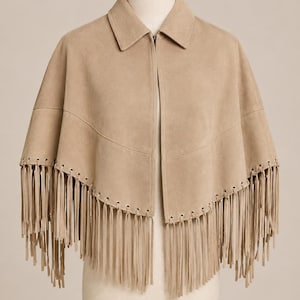 Peut inclure: Une cape en daim beige dotée d'un col classique, d'une fermeture éclair sur le devant et d'une couture horizontale sur la poitrine. L'ourlet est orné de longues franges fines fixées à travers une série de petits œillets régulièrement espacés.