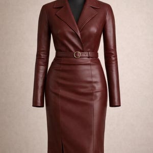 Peut inclure: Robe en cuir bordeaux à manches longues, présentée sur un mannequin. La robe a un col, une ceinture à boucle dorée et une jupe mi-longue avec une fente avant.