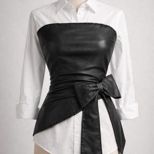 Peut inclure: Un haut de style corset en similicuir noir avec un nœud, superposé à une chemise blanche boutonnée. Le corset s'enroule autour de la taille et présente un nœud sur le côté. La chemise a des manches trois-quarts.