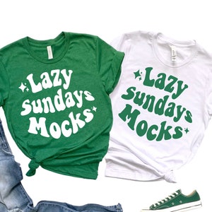 May include: Two green and white t-shirts with the text "Lazy Sundays Mocs" in a retro font. The shirts are laid out on a white background with a pair of green sneakers and blue jeans.
